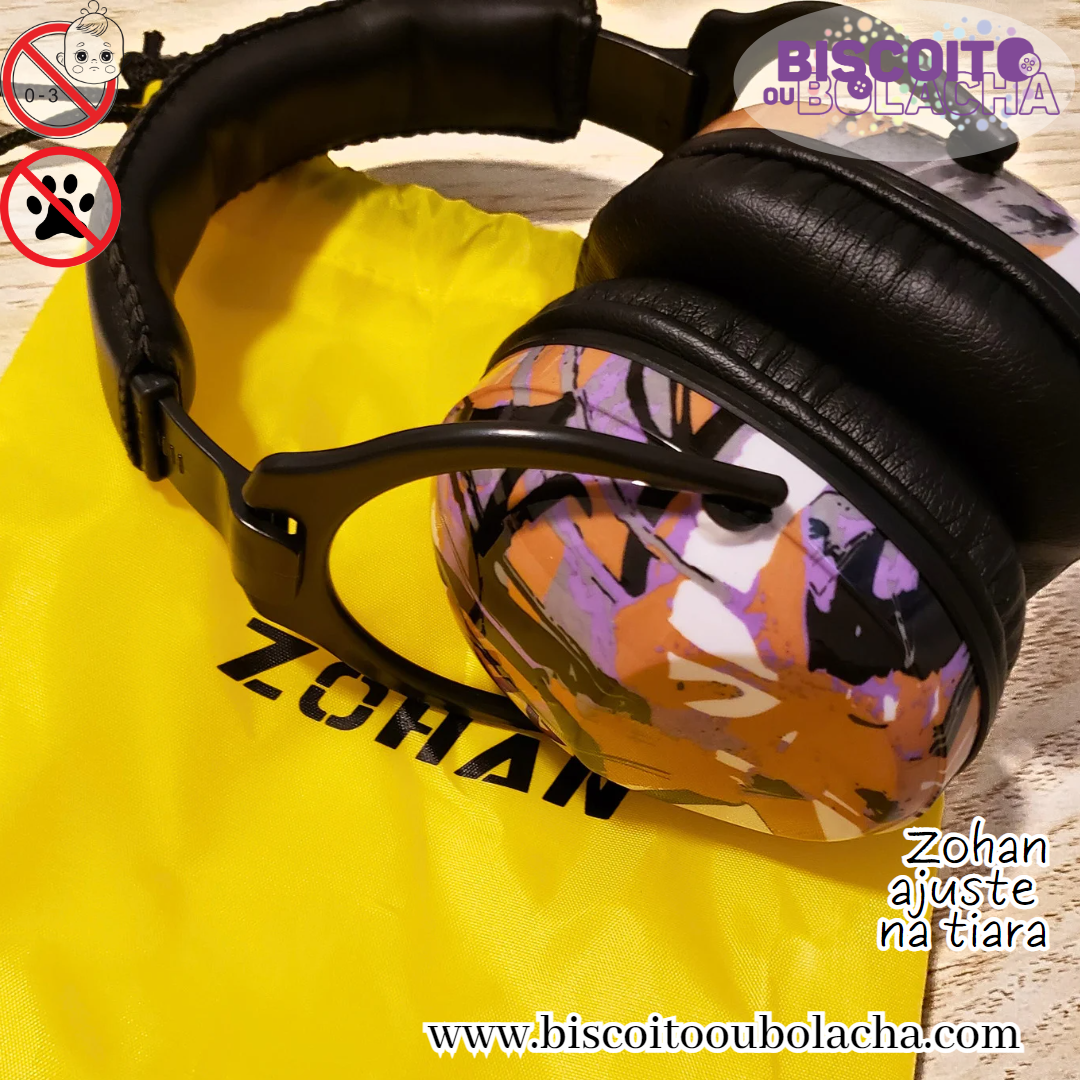 na imagem temos o abafador modelo ajuste na tiara da marca Zohan, com estampa em mix de cores mel, roxo, preto e branco na concha, sobre uma sacola de transporte de nylon na cor amarela com Zohan estampado em preto, e cordão preto no topo para fechar. Todas as conexões do abafador são de material plástico na cor preta, a capa acolchoada ao redor da tiara e a almofada da concha são em couro na cor preta. A estampa em mix de cores consiste em manchas irregulares e sobrepostas nas cores mel, roxo, preto e branco.